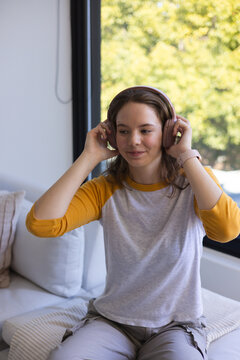 Female adjusting pink headphones while sitting on couch by window wearing smartwatch and gray tee