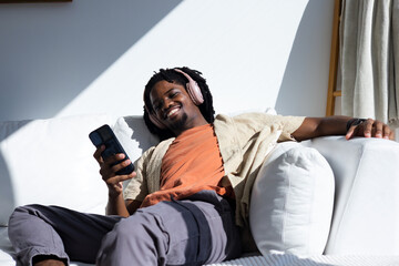 Fototapeta premium African American man reclining on white sofa at home, holding smartphone wearing pink headphones