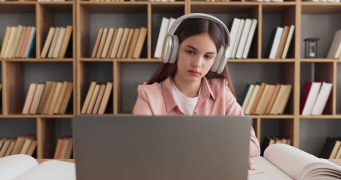 Student teenager girl with headphones studying laptop. In a quiet library she researches notes from open books and attends online learning. Best for learning. Concept of focus and concentration.