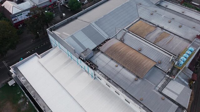 Aerial drone view of a dense industrial area featuring large warehouse buildings, factory rooftops, surrounding roads, vehicles, trees, and urban infrastructure. The composition highlights the contras