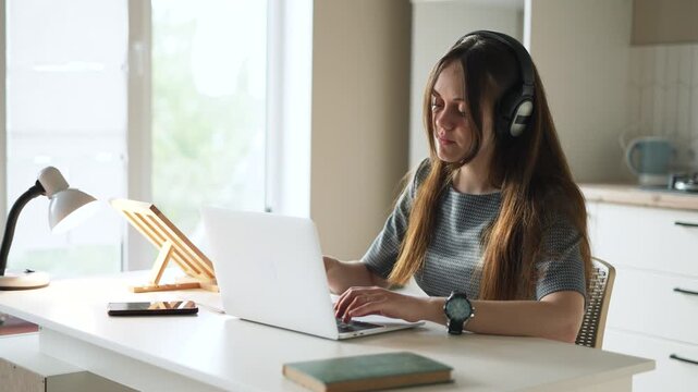 Young woman types on laptop near bright window. She smiles working from home office. Long hair frames smiling face. She enjoys remote work, online learning and video chat on laptop in sunlight.
