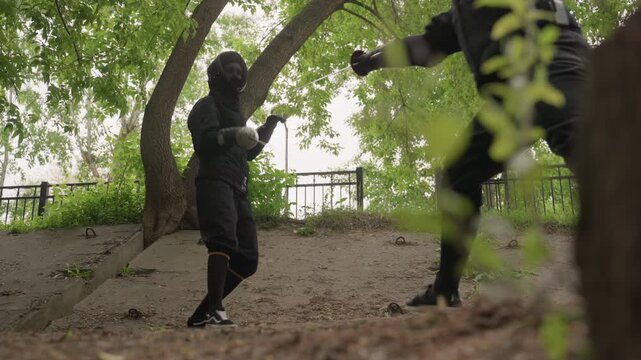 Two people in black fencing gear engage with swords outdoors under trees demonstrating techniques. One person holds sword while other moves into position both wear protective masks