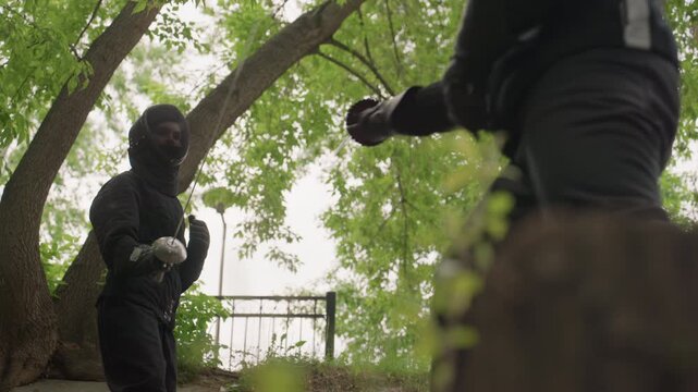 Two people in black fencing gear engage with swords outdoors under large trees demonstrating controlled movements and techniques. scene features natural greenery as backdrop while fencers