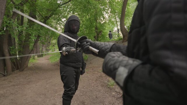 Close up of two people in black fencing gear practicing swordplay on dirt path surrounded by green trees. One person holds camera while their opponent demonstrates various fencing techniques