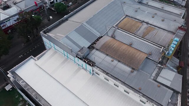 Aerial drone view of a dense industrial area featuring large warehouse buildings, factory rooftops, surrounding roads, vehicles, trees, and urban infrastructure. The composition highlights the contras