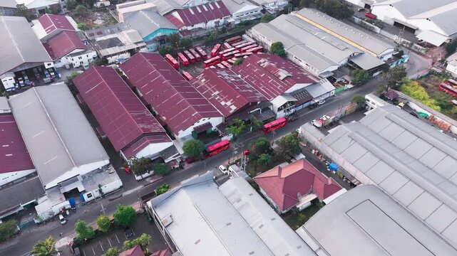 Aerial drone view of a dense industrial area featuring large warehouse buildings, factory rooftops, surrounding roads, vehicles, trees, and urban infrastructure. The composition highlights the contras
