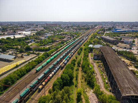 High angle aerial shot captures many long cargo trains on multiple parallel tracks beside old industrial warehouse building and green trees under clear sunny sky