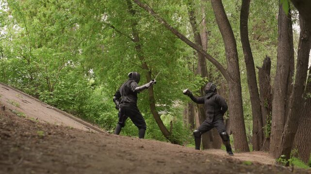 Two men in black ninja attire engage in sword fight on outdoor dirt path surrounded by dense green foliage and tall trees. Their movements are fluid as they wield swords demonstrating