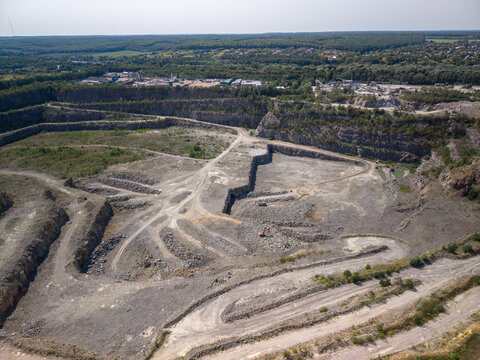 Wide drone shot captures massive industrial stone quarry with rocky terraces, winding dirt tracks, excavator machines, and mounds of gravel near forest under clear sky