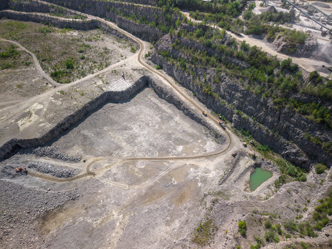 Aerial shot captures active stone quarry showing deep rocky terraces, winding transport roads with trucks, excavator machine, and small green water pond under bright daylight