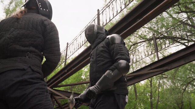 Two people in black protective gear engage in sword fight under metal beams with trees visible. One person wields sword while other prepares to defend both wearing padded clothing