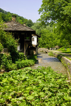 A picturesque scene of an old watermill beside a flowing stream, with a stone bridge in the background and lush green trees surrounding the area.