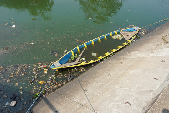 Abandoned boat rests beside muddy water. Greens and yellows stain its hull with decay. Leaves float, weeds cling to its edges. Concrete dock holds it still, unused. Polluted river reflects broken sky
