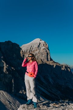 Woman in a pink top shades her eyes on a rocky slope below a pointed Dolomite summit. Sun protection and active mountain travel for eyewear, sportswear and outdoor tourism marketing