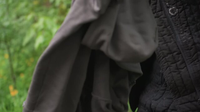 Close up of man putting on and storing clothing in outdoor setting. man handles various garments including jackets or coats while interacting with bags and backpacks. Background features