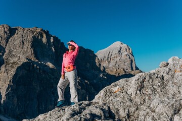 Woman in a pink top stands on a rocky ridge with a hand shading her eyes from the sun. Freedom and confidence of solo mountain hiking for travel, wellness and sportswear brands © Екатерина Лукашина