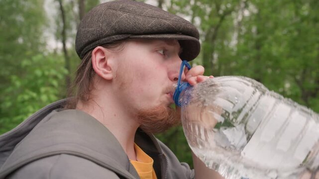 Close up of man drinking water from large plastic bottle in outdoor park setting. Man holds clear condensation covered container to mouth while wearing flat cap and jacket background shows