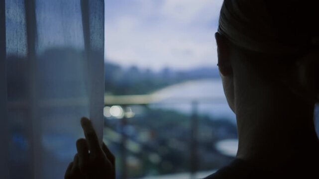 Blonde woman looking at window from hotel room, back view, wait and feel nervous . Relax time in summer vacation, lady touching white curtain, enjoy window view on garden and territory of luxury hotel