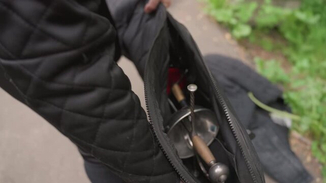 Close up of person in black attire holding open bag containing fencing equipment revealing swords and protective gear. scene shows detailed texture of metal blades with wooden handles