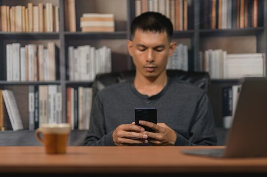 Young Man Sitting at Desk Using Smartphone in Modern Office with Bookshelf