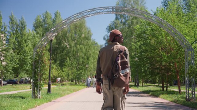 Man wearing brown jacket and cap walks down paved path in outdoor park carrying backpack. scene features green trees lining pathway under clear blue skies with sunlight casting shadows