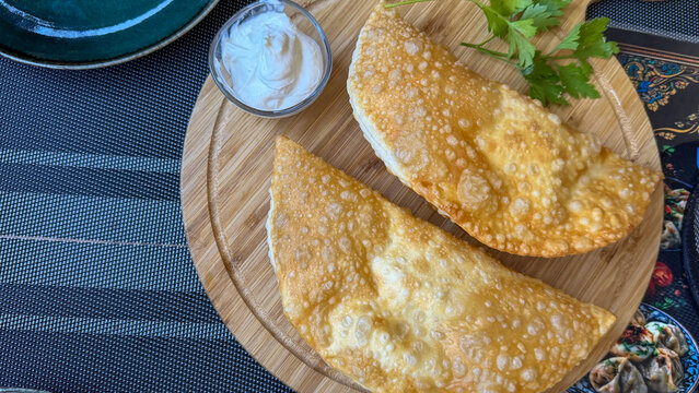 Traditional crispy chebureki, deep-fried meat-filled pastries served with sour cream and fresh parsley on a wooden board.