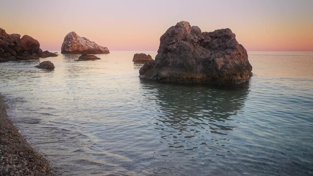 Slow motion of gentle sea waves rippling on a pebble beach near massive coastal rocks during sunset. Meditative water footage serves premium hotel website headers or relaxation vlogs as a design eleme