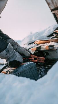 Winter car battery repair with gloved hands connecting booster cable under open hood during snowy roadside emergency in cold mountain weather