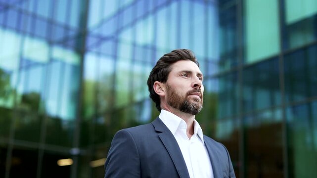Serious mature businessman walks outside office building, standing in close up. Professional in formal suit confidently looking around with a composed, accomplished expression, success, determination.