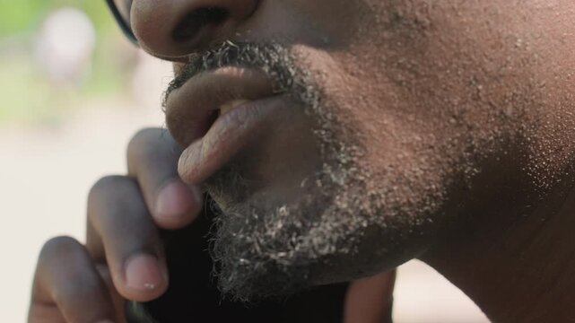 Close up of man with facial hair holding and using smartphone showing natural skin texture details. subject hand grips device while his mouth moves as if speaking or listening. Soft outdoor