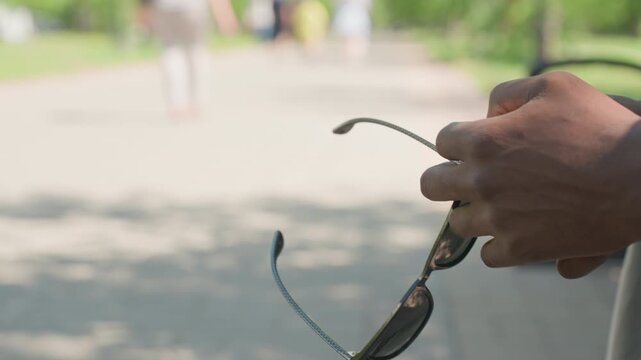Close up of hand holding sunglasses with blue patterned arms in outdoor park setting. person holds glasses steadily showcasing their design against soft natural light and blurred greenery