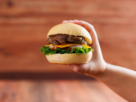 Delicious cheeseburger layer with golden bun juicy meat beef or pork patty melted cheddar cheese holding by hand on wooden background fresh ingredients lettuce tomatoes tasty classic burger junk food.