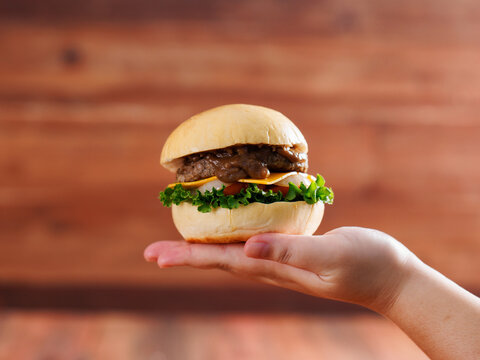 Delicious cheeseburger layer with golden bun juicy meat beef or pork patty melted cheddar cheese holding by hand on wooden background fresh ingredients lettuce tomatoes tasty classic burger junk food.