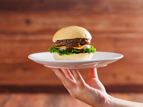Hand holding delicious cheeseburger on plate layer with golden bun juicy meat beef or pork patty melted cheddar cheese on wooden background fresh ingredients lettuce tomatoes tasty burger junk food.