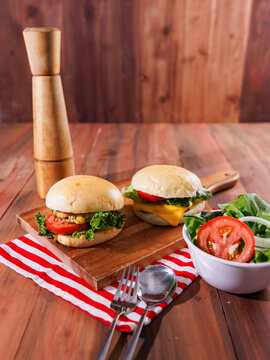 Two delicious homemade cheeseburgers with fresh lettuce tomatoes served on wooden board with salad wooden pepper grinder spoon fork on wood table with red striped cloth burger fast food advertising.