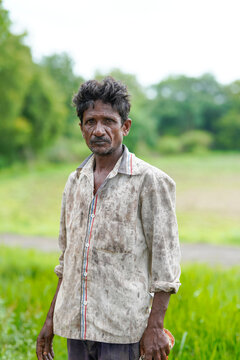 a hard-working Indian farmer standing outdoors in a field wearing a mud-stained shirt after a long day of labor.