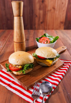 Two delicious homemade cheeseburgers with fresh lettuce tomatoes served on wooden board with salad wooden pepper grinder spoon fork on wood table with red striped cloth burger fast food advertising.