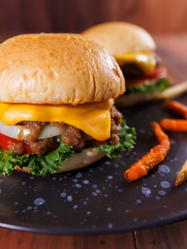 Close up of juicy cheeseburger top melted cheese beef pork patty lettuce tomato onion served with crispy french fries on black plate warm rustic background dining tasty classic fast food advertising.