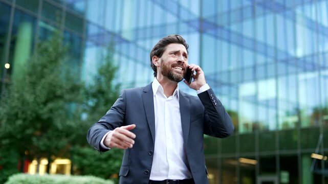 Furious manager in formal suit shouting aggressively while talking on mobile phone outside modern glass office building. Angry businessman screaming during stressful business phone call conversation.