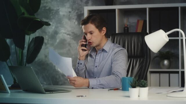 Annoyed young CEO talks on phone while reviewing failed business deals in office