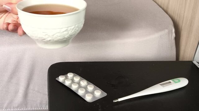 Sick woman putting a cup of hot tea beside a thermometer, seasonal flu, respiratory infections, winter viruses and high illness rates during the cold season.