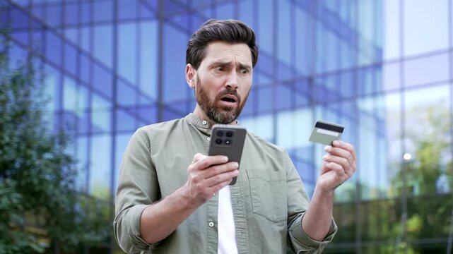 Frustrated businessman holding credit card and mobile phone in shock after losing money to online deception near glass office building. Employee discovering bank fraud and stolen account balance.