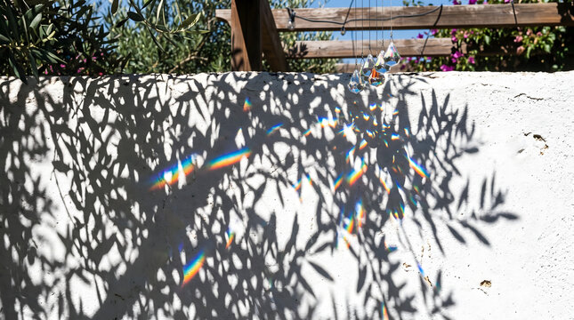 Beautiful rainbow light prism reflections and dark shadows of olive tree leaves cast on a rustic white stucco wall in a sunny Mediterranean garden during a warm and bright summer day