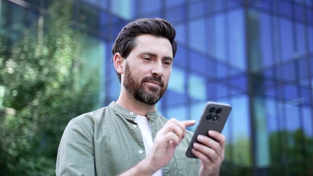 Mature businessman checking information on smartphone standing outside modern office building on city street. Professional reviewing mobile content during workday in urban business district. Close up
