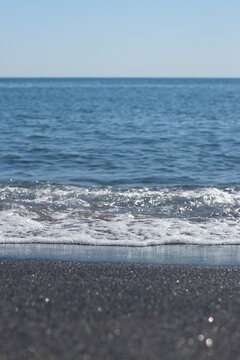 Calm blue sea under clear sky on a sunny day. Sea wave with white foam approaching sandy shore. Aesthetic summer landscape with sea waves and horizon. Close-up of sea surf on dark sand