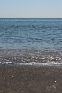 Calm blue sea under clear sky on a sunny day. Sea wave with white foam approaching sandy shore. Aesthetic summer landscape with sea waves and horizon. Close-up of sea surf on dark sand