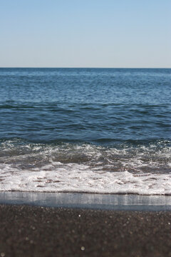 Calm blue sea under clear sky on a sunny day. Sea wave with white foam approaching sandy shore. Aesthetic summer landscape with sea waves and horizon. Close-up of sea surf on dark sand