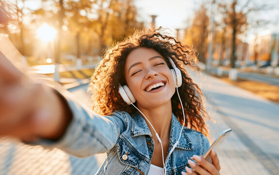 A smiling young woman with curly hair, wearing headphones and a denim jacket, taking a selfie in a park on a sunny day, holding her phone and looking at the camera with a joyful expression
