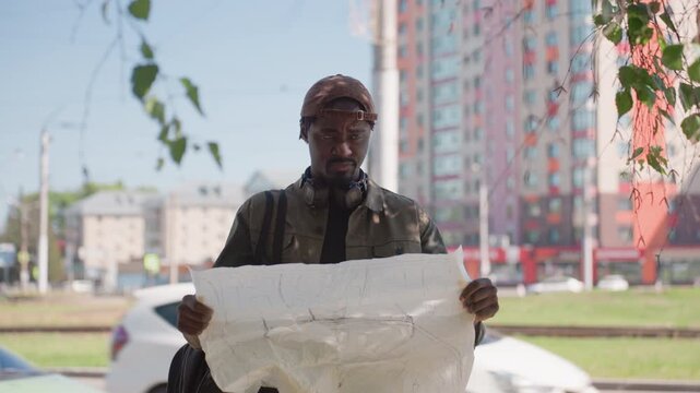 Close up of man holding large hand drawn map in urban setting with tall buildings and passing cars. Man wears casual outdoor clothing including cap jacket headphones around neck carries bag