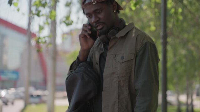 Close up of man holding smartphone and later speaking on it while standing outdoors with bag. Man interacts with crumpled piece of paper wearing headphones around neck in casual attire